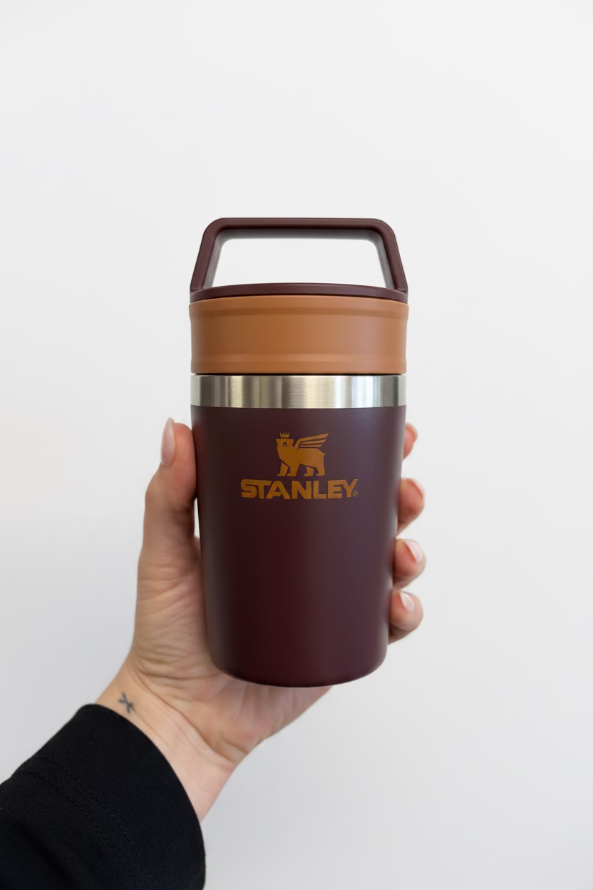 A hand holding a Stanley vacuum-insulated tumbler with a dark plum body, a tan lid, and a stainless steel band, against a textured off-white wall.